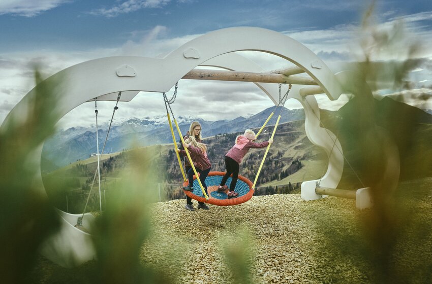 Kind schaukelt auf großer Netzschaukel mit Mama vor Bergkulisse, moderne Spielplatzstruktur in Naturumgebung. | © Grossarler Bergbahnen GmbH & Co KG
