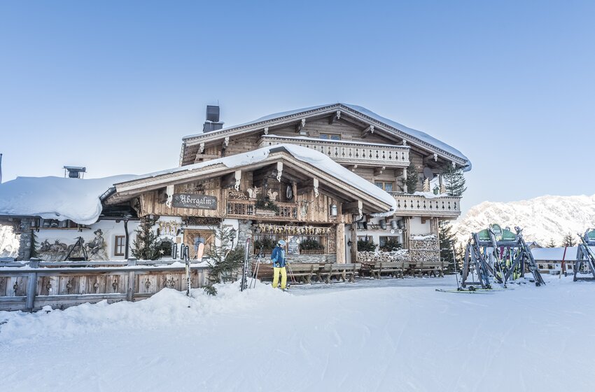 Verschneite Abergalm mit Skifahrer, Skiern und Holzbänken vor schneebedeckten Bergen unter blauem Himmel | © Creatina Werbedesign & Photography
