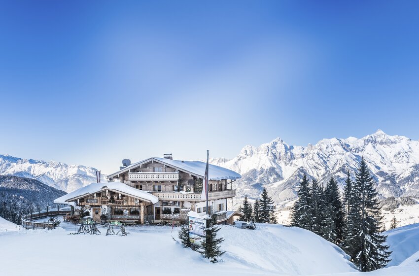 Verschneite Berghütte mit Terrasse und Tannen vor einem weitläufigen Panorama schneebedeckter Alpengipfel | © Creatina Werbedesign & Photography