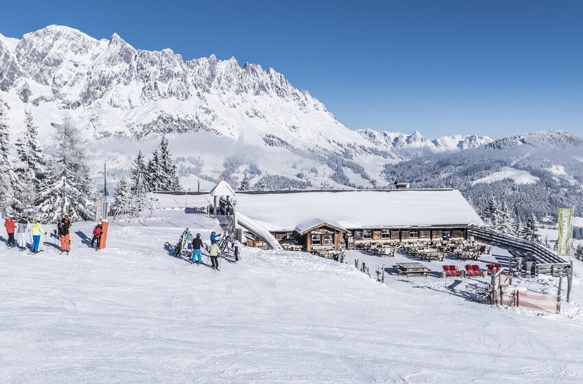 Karbachalm mit Terrasse an breiter Skipiste, umgeben von Skifahrern und Skiständern vor schneebedeckten Bergen | © Creatina Werbedesign & Photography