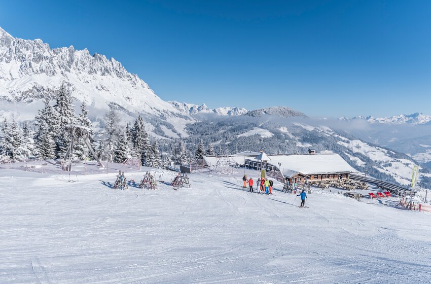 Karbachalm an breiter Skipiste mit Skifahrern und Skiständern vor verschneiten Bergen unter blauem Himmel | © Creatina Werbedesign & Photography
