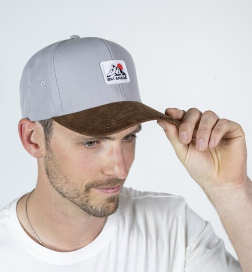 Mann trägt graues Baseballcap mit Ski amadé Logo und braunem Schirm und hält den Schirm vor hellem Studiohintergrund | © Ski amadé GmbH / Lorenz Masser