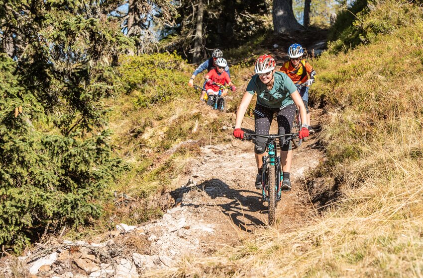 Gruppe mit Erwachsenen und Kindern fährt mit Mountainbikes auf schmalem Wald-Trail durch sonnige Berglandschaft. | © Reiteralm Bergbahnen GmbH & Co.KG