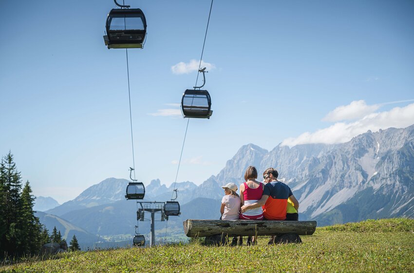 Familie sitzt auf einer Bank mit Blick auf Gondeln und das imposante Dachsteinmassiv. | © Ski amadé GmbH