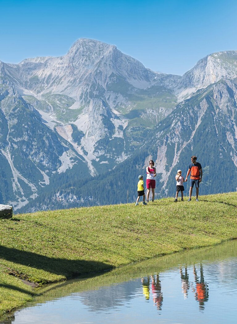 Familie wandert entlang eines klaren Sees mit Bergpanorama und spiegelnder Wasseroberfläche. | © Ski amadé GmbH